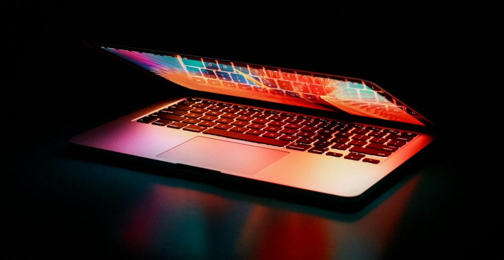 Laptop partially opened, emitting vibrant multicolored lights in dark setting.