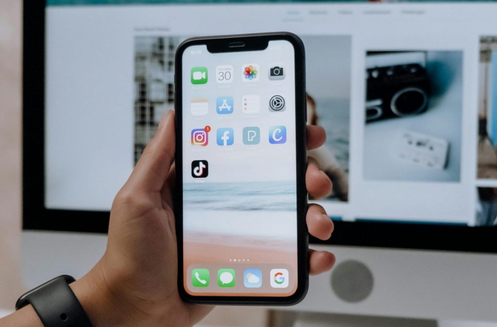 Close-up of a hand holding a smartphone showing social media apps in front of a desktop computer.